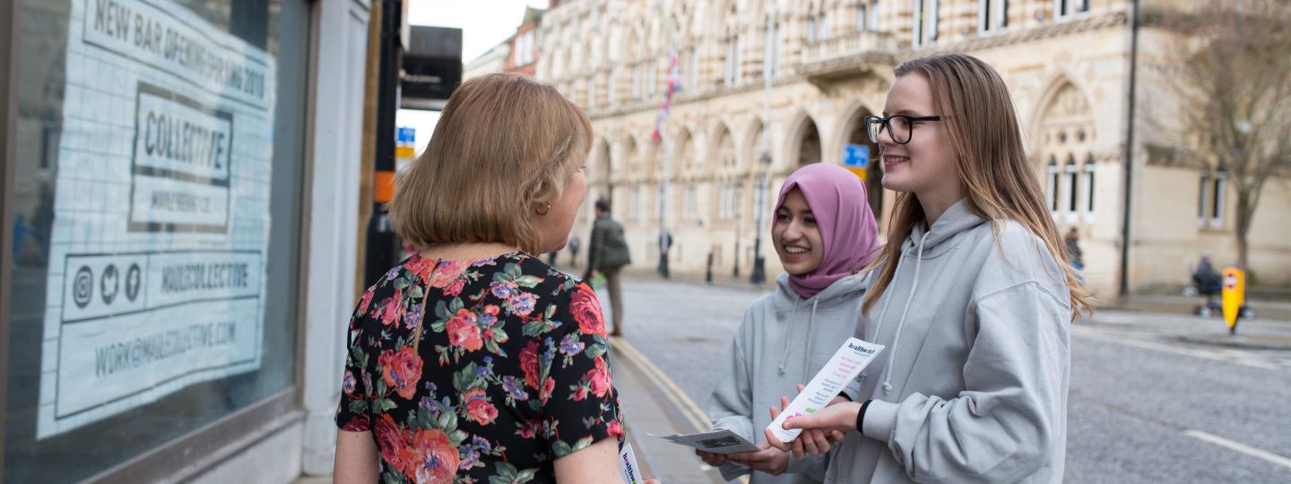Two young girls talking to a lady and showing a Healthwatch leaflet